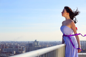 Girl brunette in blue dress with closing eyes stands on roof of tall building on wind