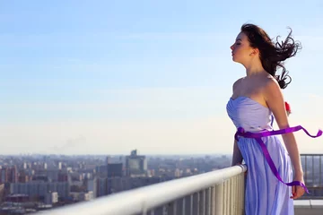 Foto op Aluminium Girl brunette in blue dress with closing eyes stands on roof of tall building on wind © Pavel Losevsky