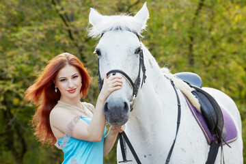 Young smiling red-haired woman holds white horse by bridle in park