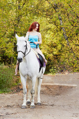Young smiling red-haired woman rides on white horse in park