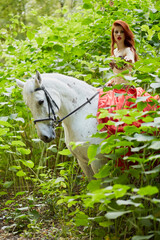 Red-haired young woman in red dress sits on white horse among green plants in park