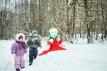 Fototapeta na wymiar Little boy and girl stand looking at stuffed dummy Maslenitsa that sits on snowbank in winter park