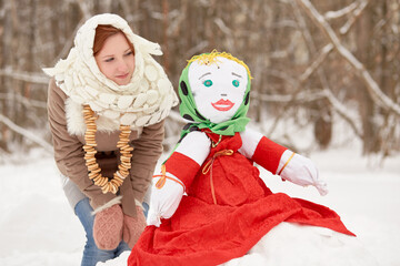 Teenage girl stands and lloks at stuffed dummy Maslenitsa, sitting on snowbank in winter park