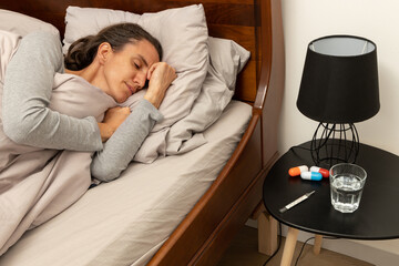 Ill young lady sleeps in bed in the day with medicine, water and thermometer on bedside cabinet;...