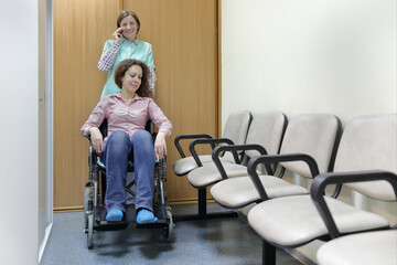 Nurse talking on phone and smile in hospital corridor, standing next to patient in wheelchair, holding handles