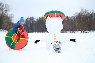Happy boy with tube stands near upside down snowman at winter day