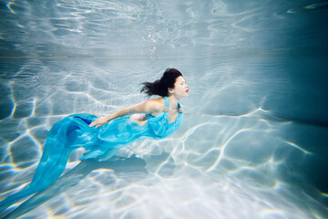 Young black-haired woman in blue dress swims underwater with her eyes close