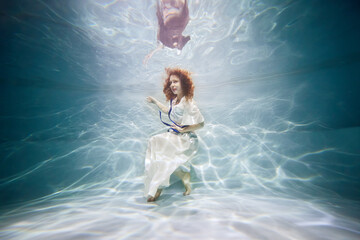 Smiling young woman in white dress poses in swimming pool underwater