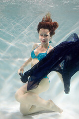 Smiling young woman in blue swimsuit poses in swimming pool with dark-blue fabric in hands underwater - obrazy, fototapety, plakaty