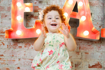 Little happy girl claps and sits near wall with big letters in studio