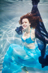 Smiling young woman in blue swimsuit poses in swimming pool with dark-blue fabric underwater - obrazy, fototapety, plakaty