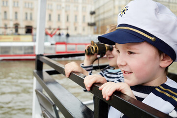 Two little boys one in cap and another with binocular stand on boat stern