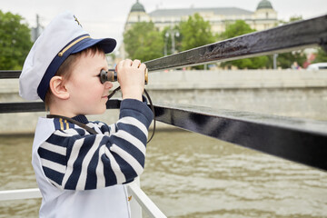 Obraz na płótnie Canvas Little boy dressed as a captain looks through binoculars standing at boat stern