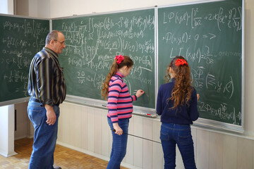 Teacher and two girls stand near blackboard with formulas in classroom, girl writes