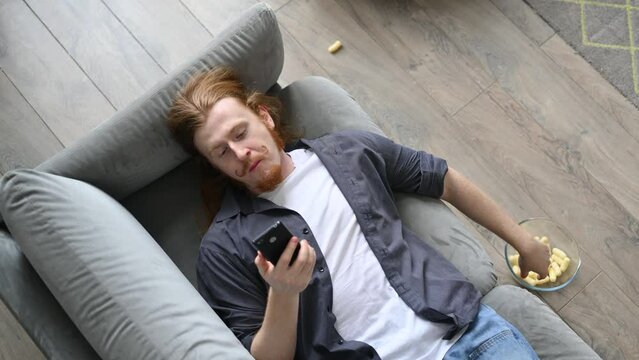 A young red-haired guy is lying on the sofa and eating corn flakes, scrolling through social networks on his phone