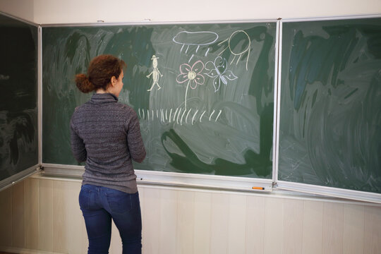 Young woman stands near blackboard with pictures in classroom, back view, focus on picture
