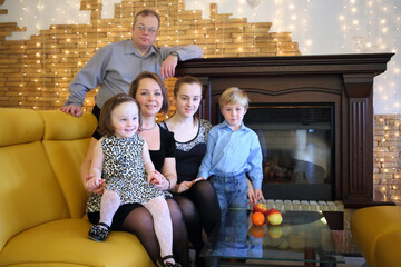 Family of five people in elegant clothes sit on a sofa near the fireplace