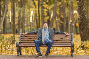 Fototapeta premium Portrait of young man in gray coat and jeans sitting on bench in alley in autumn park, hands on back, looking up
