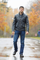Full length portrait of young dark-haired man in black jacket on avenue in park, walking