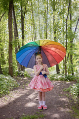 Little girl in polka-dotted dress stands with rainbow coloured umbrella in summer park