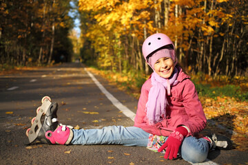 Happy girl in a protective helmet sits outstretched leg in the rollers on the asphalt in the autumn...