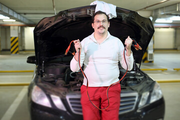 Man with wires for recharging the battery near an open hood of the car in the underground parking