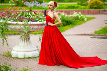 Obraz na płótnie Canvas Full portrait of beautiful woman in red long dress standing in summer park around flowerpot with flowers, look at camera
