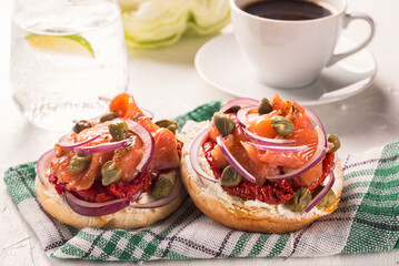 Two sandwiches with cheese cream, red fish, onions and capers on a napkin against the background of a cup of black coffee close-up.