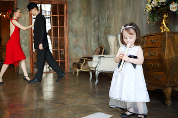 Little girl in dress plays flute in room with dancing couple, focus on child