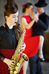 Pretty girl playing saxophone next to dancing pair, focus on musician
