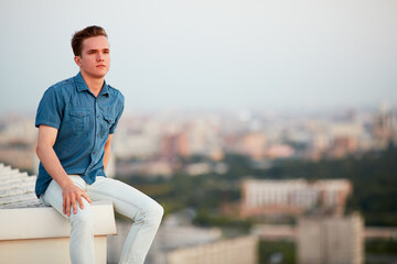 Young man in denim clothes sits on the edge of the building roof.
