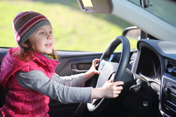 Happy little girl in pink waistcoat and striped hat sitting at wheel of car