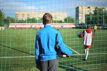 Young man in a blue sports jacket with a ball watching a football game at the stadium over the net, view from the back
