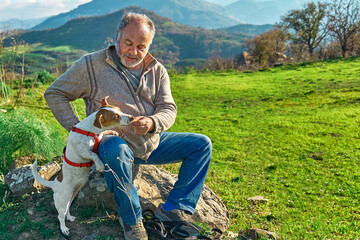 Gray haired man taking a walk with his jack russell dog in meadow in mountainous area. Mature man playing outdoors with his dog in nature.