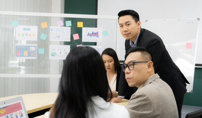 A group of people are gathered around a table with a whiteboard behind them. They are discussing something important, and one of the men is wearing glasses