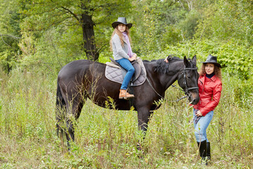 Woman in red jacket holds by the bridle horse on whom her daughter sits. Both in cowboy hats.