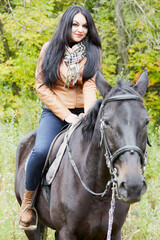 Black-haired woman in brown jacket sits on bay horse in park.