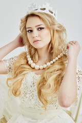 Young blond woman in crown holds beads of big artificial pearls around her neck.