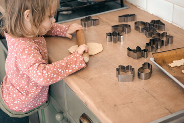 3-year-old girl standing on a stool, rolling out cookie dough with a wooden rolling pin on the...