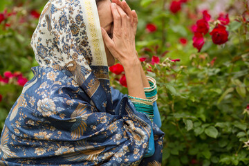Woman in beautiful sari and necklace prays with closed face among rose bushes in park - obrazy, fototapety, plakaty