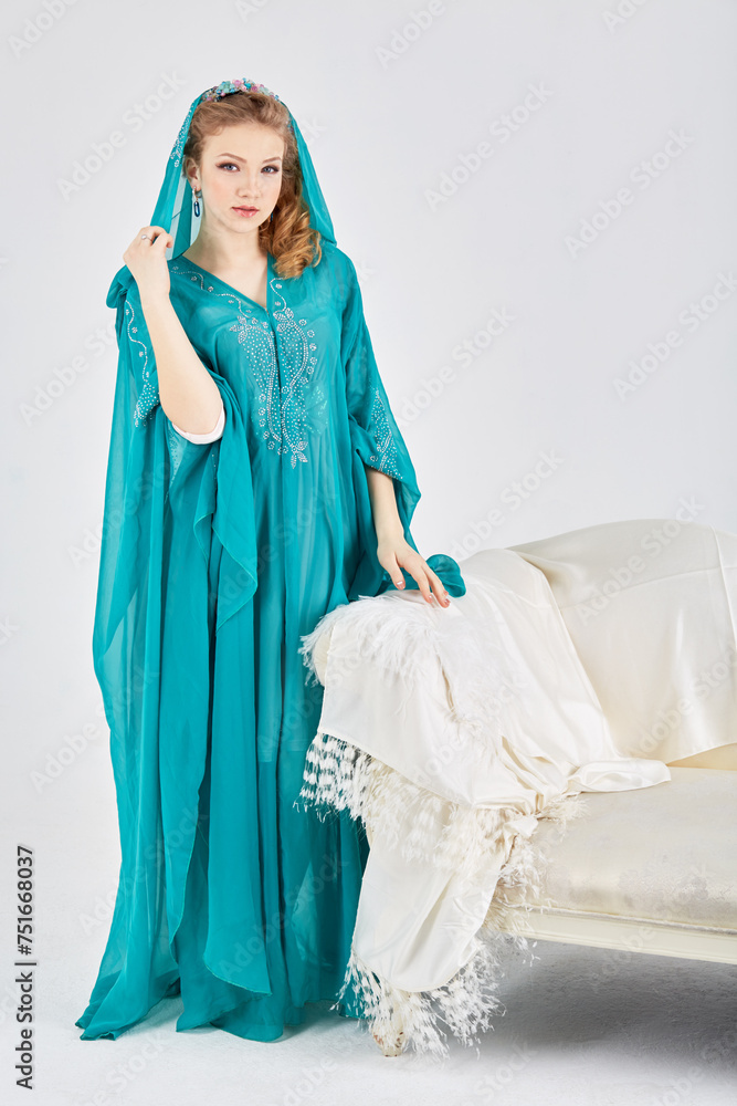 Wall mural Young woman in long dress with hood stands near couch in studio.