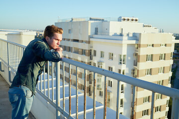 Young man in denim clothes stands on the building roof leaning to the fencing