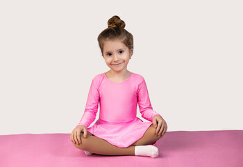 Little blonde girl in pink dress sitting on a fitness mat with her legs crossed and looking at the camera with a smile on a white background.