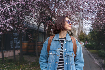 Young Caucasian happy cheerful woman in sunglasses walking through a spring city with blooming tree - 751670246