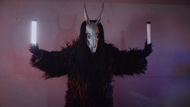 The devil monster dances with RGB sticks in his hands. A mystical man in a scary mask in a dark studio. The man is dressed in a black coat and a mask with horns on his head. Alien from space. Horror,