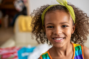 Curly-haired mixed race girl smiling, in a colorful striped dress and lime headband, captures a cheerful demeanor. Perfect for family, sibling, joy, and multicultural themes.