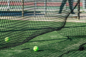 moment on a padel court, focusing on a bright green padel ball that stands out against the artificial grass surface. A black net, slightly lifted from the ground, adds depth and texture to the scene.