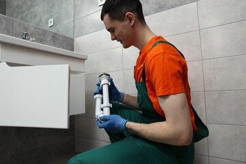 Young plumber wearing protective gloves repairing sink in bathroom