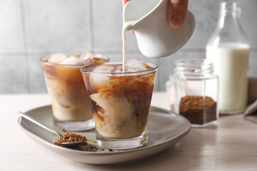 Woman pouring milk into glass with refreshing iced coffee at light table, closeup
