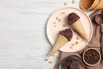 Chocolate ice cream scoops in wafer cones and candies on light wooden table, flat lay. Space for...
